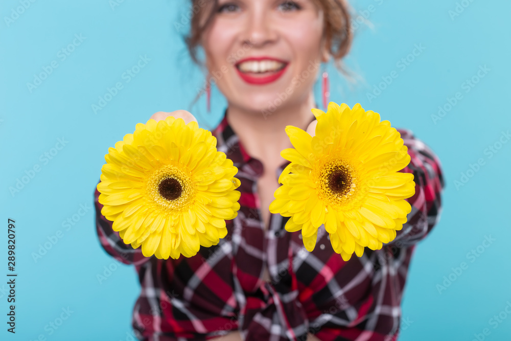 Portrait of positive young woman in plaid shirt holding two yellow flowers with different hearts near breast posing against blue background. Concept of body beauty in all forms, Body positivity