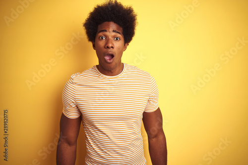 American man with afro hair wearing striped t-shirt standing over isolated yellow background afraid and shocked with surprise and amazed expression, fear and excited face.