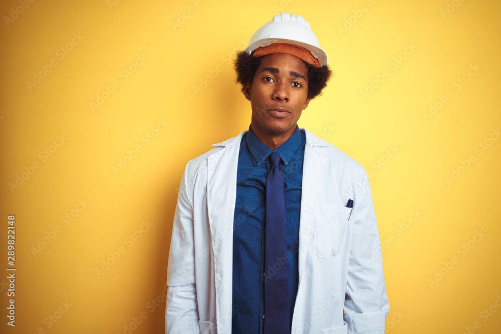 Afro american engineer man wearing white coat and helmet over isolated yellow background with serious expression on face. Simple and natural looking at the camera.