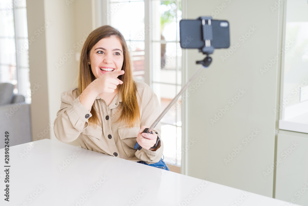 Beautiful young woman taking a picture using selfie stick very happy pointing with hand and finger Beautiful young woman taking a picture using selfie stick very happy pointing with hand and finger