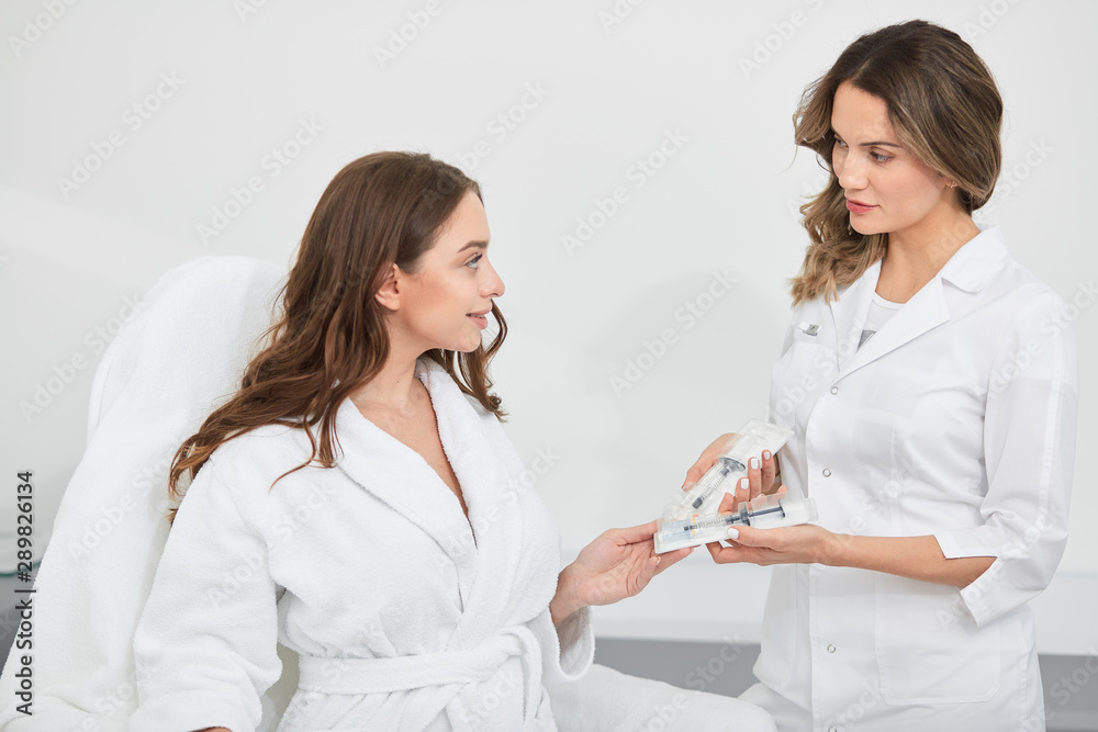 beautiful doctor holding two syringe, offering them to a clien in white robe sitting in the armchair. close up photo. lifestyle . business beautiful doctor holding two syringe, offering them to a clien in white robe sitting in the armchair. close up photo. lifestyle . business