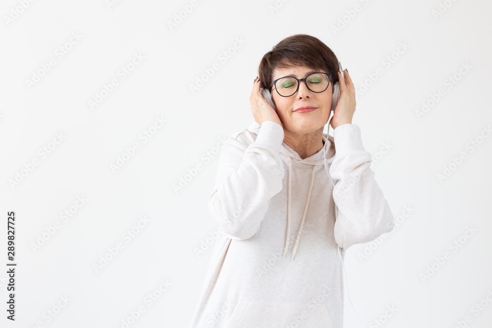 Cute positive middle-aged woman in sweater and glasses is listening to music with wire headphones standing on a white background with copy space. Concept of hobbies and subscriptions to favorite radio Cute positive middle-aged woman in sweater and glasses is listening to music with wire headphones standing on a white background with copy space. Concept of hobbies and subscriptions to favorite radio