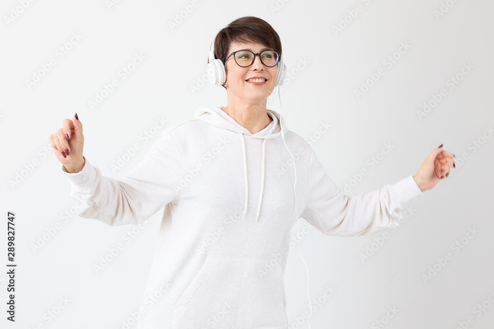 Pretty keen middle-aged woman in glasses and a white sweater listens to her favorite music with headphones on a white background. Online radio and music subscription concept. Pretty keen middle-aged woman in glasses and a white sweater listens to her favorite music with headphones on a white background. Online radio and music subscription concept.