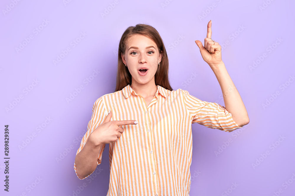 Lovely female model telling funny story, trying not burst into laugh,  pointing up with one hand, and to the side with another, looking in camera,  has cute face expression. Stock Photo |