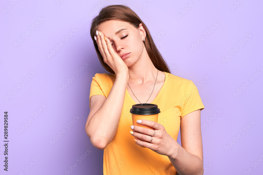 Studio shot of sleepy female student holding paper cup of coffee to go, standing with closed eyes, with one hand on her face, extremely tired after sleepless night, prepared to difficult exam. Studio shot of sleepy female student holding paper cup of coffee to go, standing with closed eyes, with one hand on her face, extremely tired after sleepless night, prepared to difficult exam.