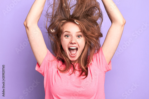 Close up portrait shot of playful young female jumping high, shouting loudly, messy hair upwards, suprised and happy to get present, dreams come true, birthday girl, modeling over light purple wall.