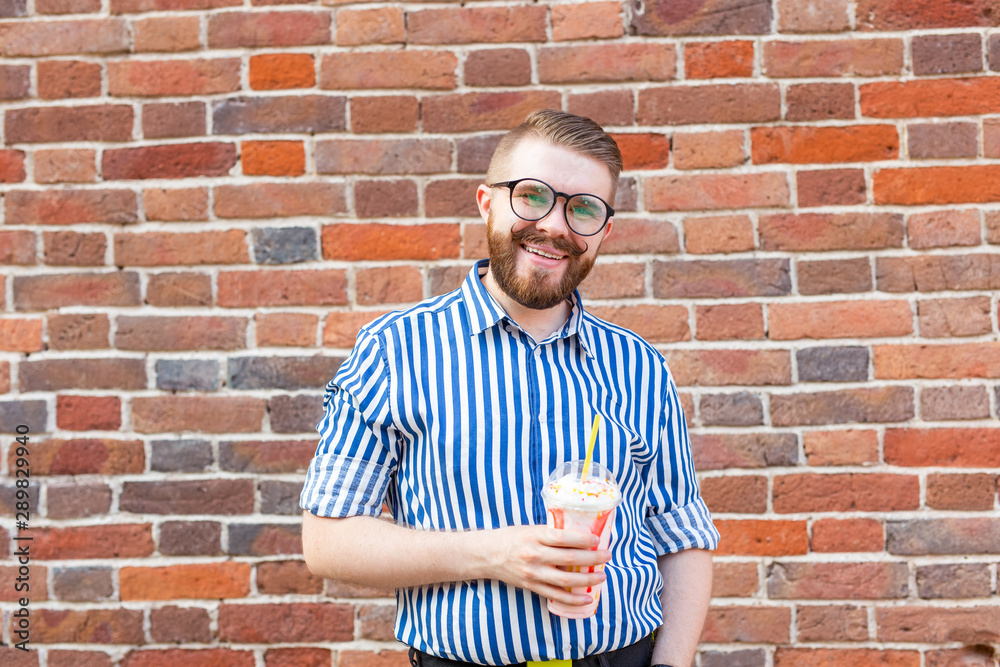 Blurred positive young hipster man with beard and mustache in glasses holding a fruit milkshake in his hands posing on the street near a brick wall. Concept of summer walks. Blurred positive young hipster man with beard and mustache in glasses holding a fruit milkshake in his hands posing on the street near a brick wall. Concept of summer walks.