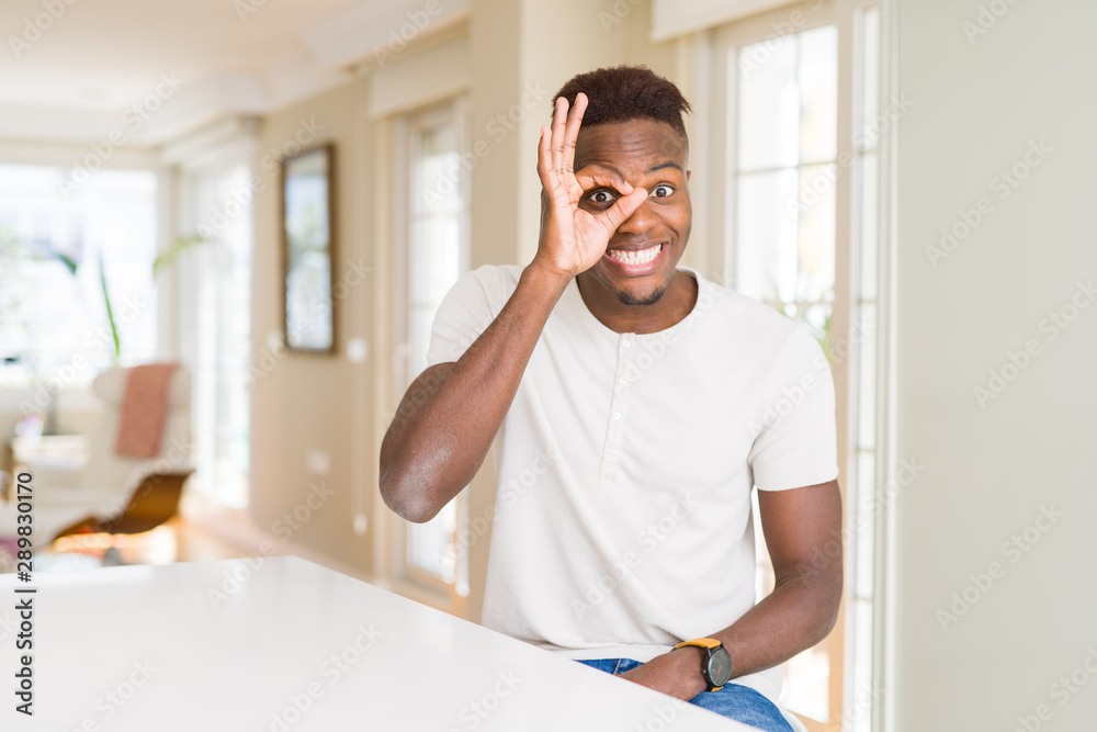 Handsome african american man on white table at home doing ok gesture with hand smiling, eye looking through fingers with happy face. Handsome african american man on white table at home doing ok gesture with hand smiling, eye looking through fingers with happy face.