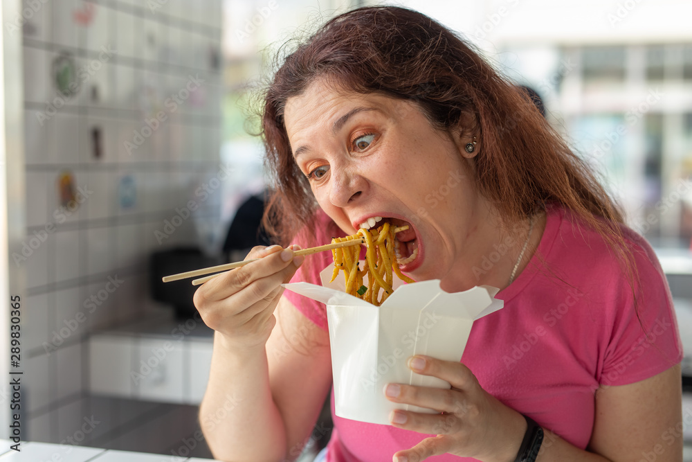 Close-up portrait of an amusing crazy young pretty girl eating chinese noodles with wooden chopsticks sitting in a cafe. Concept of hunger and satiety. Close-up portrait of an amusing crazy young pretty girl eating chinese noodles with wooden chopsticks sitting in a cafe. Concept of hunger and satiety.