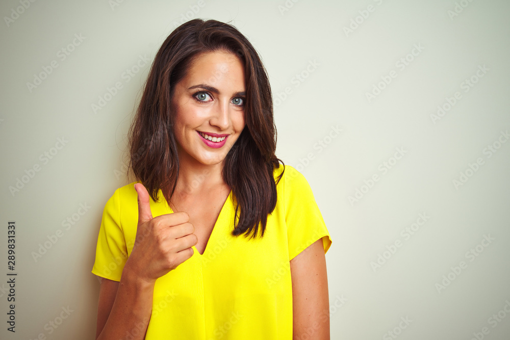 Young beautiful woman wearing yellow t-shirt standing over white isolated background doing happy thumbs up gesture with hand. Approving expression looking at the camera with showing success. Young beautiful woman wearing yellow t-shirt standing over white isolated background doing happy thumbs up gesture with hand. Approving expression looking at the camera with showing success.
