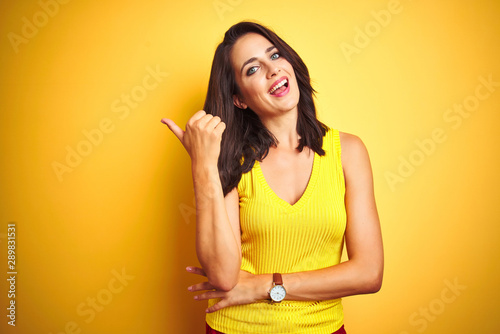 Young beautiful woman wearing t-shirt standing over yellow isolated background smiling with happy face looking and pointing to the side with thumb up.