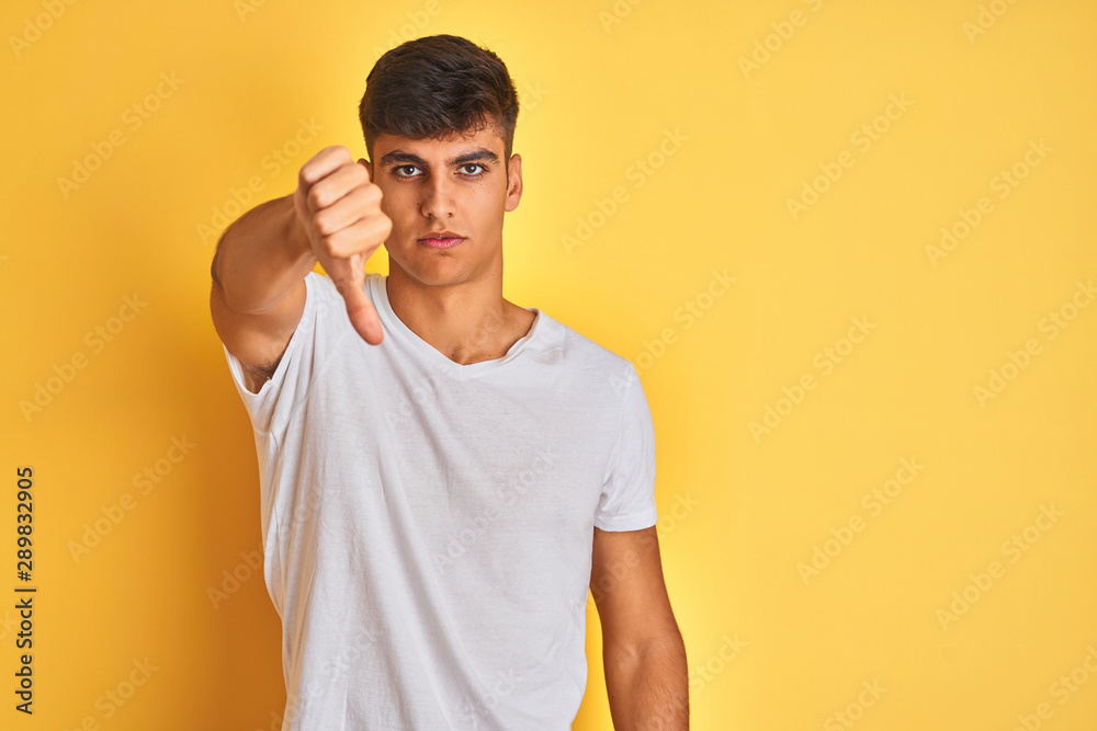 Young indian man wearing white t-shirt standing over isolated yellow background looking unhappy and angry showing rejection and negative with thumbs down gesture. Bad expression. Young indian man wearing white t-shirt standing over isolated yellow background looking unhappy and angry showing rejection and negative with thumbs down gesture. Bad expression.