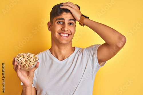 Young indian man holding bowl with pistachios over isolated yellow background stressed with hand on head, shocked with shame and surprise face, angry and frustrated. Fear and upset for mistake.