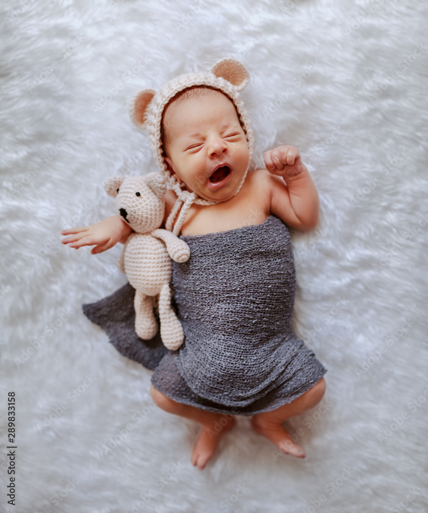 Top view of sleepy newborn boy wrapped in gray scarf and with little cap hugging knitted bear and yawning. - obrazy, fototapety, plakaty 