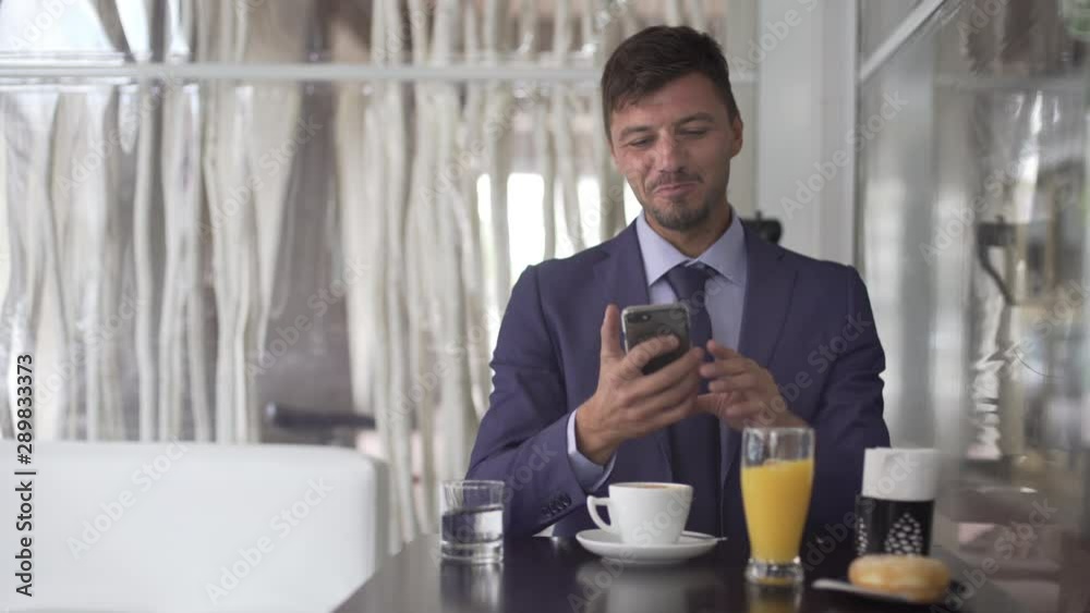 Attractive young bearded happy businessman at the coffee shop using smartphone, drinking a cup of coffee, orange juice and eating a doughnut. Expressions of a business man. Attractive young bearded happy businessman at the coffee shop using smartphone, drinking a cup of coffee, orange juice and eating a doughnut. Expressions of a business man.