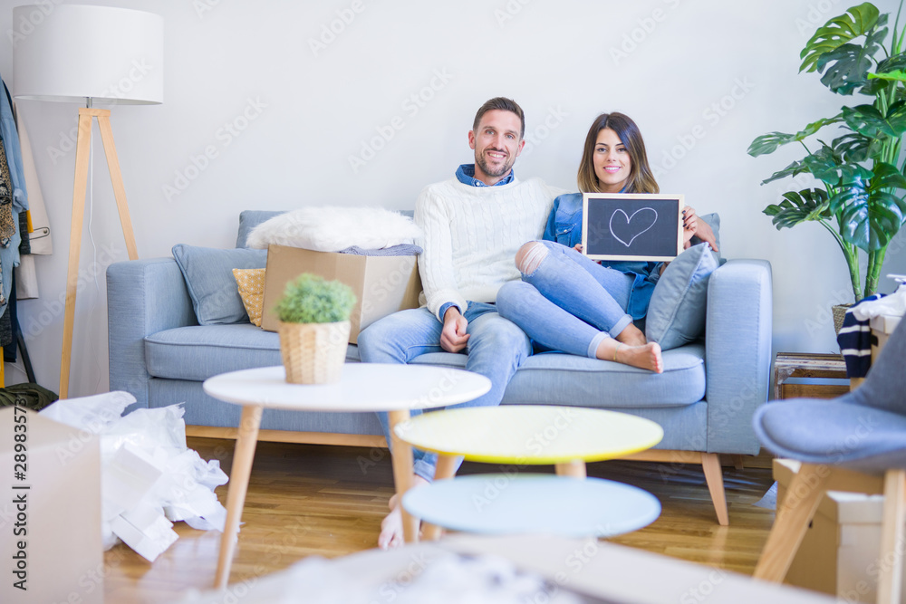 Young beautiful couple sitting on the sofa holding blackboard with a heart at new home around cardboard boxes Young beautiful couple sitting on the sofa holding blackboard with a heart at new home around cardboard boxes
