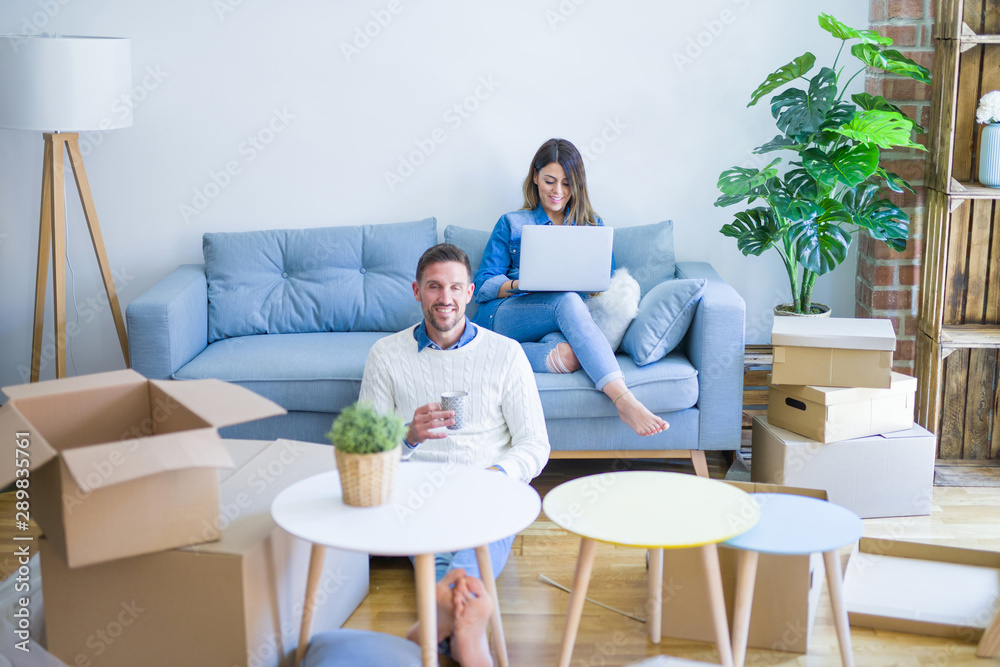 Young beautiful couple sitting on the sofa drinking cup of coffee using laptop at new home around cardboard boxes Young beautiful couple sitting on the sofa drinking cup of coffee using laptop at new home around cardboard boxes