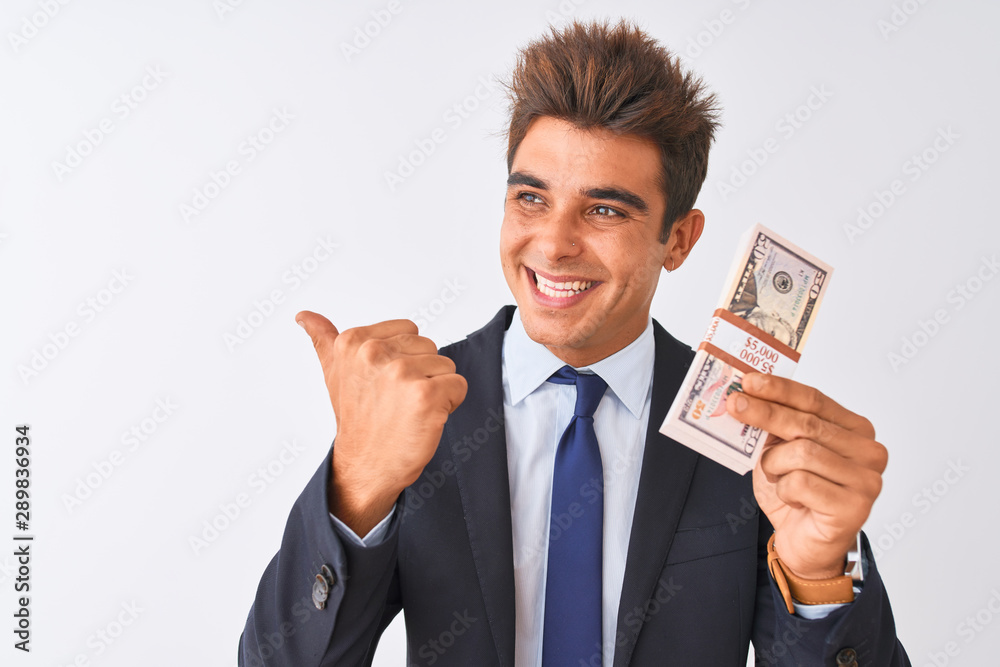 Young handsome businessman wearing suit holding dollars over isolated white background pointing and showing with thumb up to the side with happy face smiling Young handsome businessman wearing suit holding dollars over isolated white background pointing and showing with thumb up to the side with happy face smiling