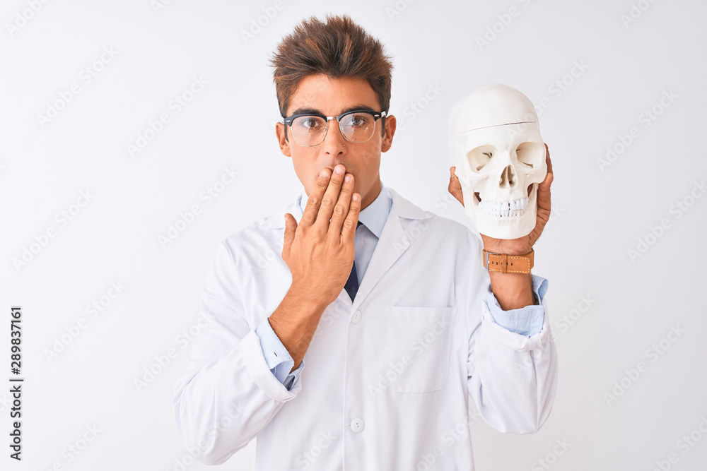 Young handsome doctor man wearing glasses holding skull over isolated white background cover mouth with hand shocked with shame for mistake, expression of fear, scared in silence, secret concept Young handsome doctor man wearing glasses holding skull over isolated white background cover mouth with hand shocked with shame for mistake, expression of fear, scared in silence, secret concept