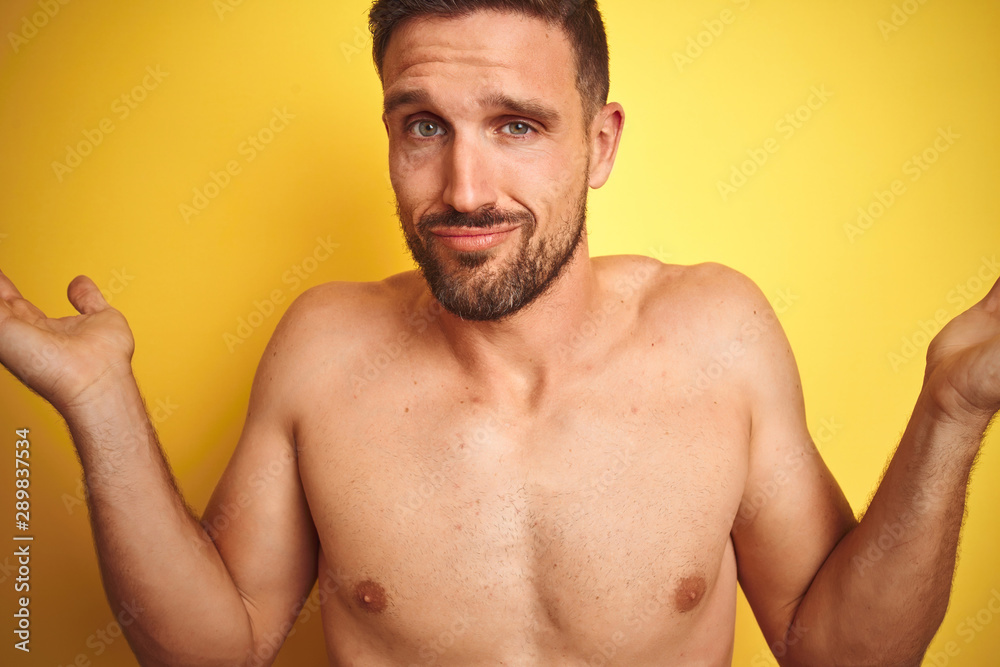 Fototapeta premium Sexy young shirtless man showing nude fitness chest over yellow isolated background clueless and confused expression with arms and hands raised. Doubt concept.