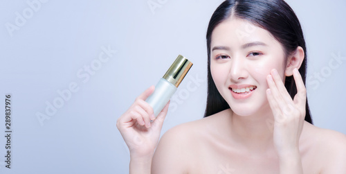 Beauty treatment. Asian woman applying moisturizing cream on face, holding bottle with skin care product, studio shot on blue background.