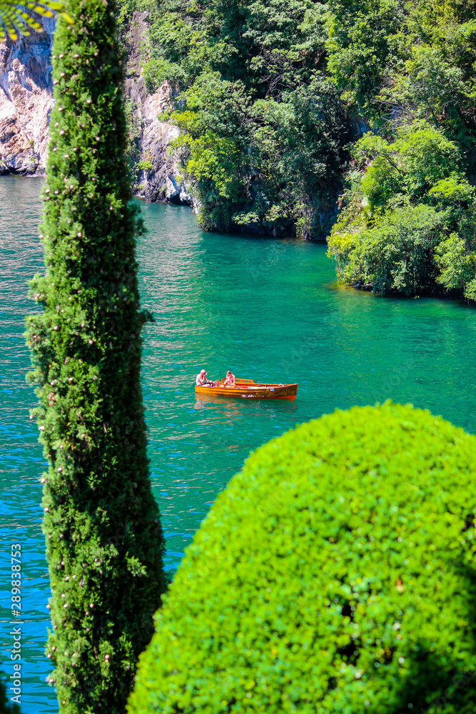a wooden boat near the Balbianello villa with trees in the foreground. The Villa del Balbianello is a villa in the comune of Lenno, Italy, overlooking Lake Como.