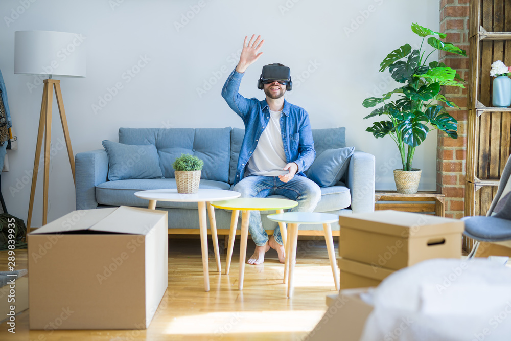 Young man wearing virtual reality glasses playing a simulation game sitting on the sofa around cardboard boxes moving to a new house