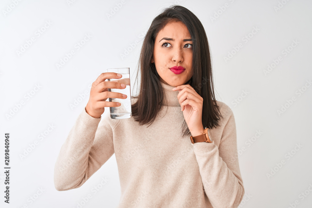 Young beautiful chinese woman holding glass of water standing over isolated white background serious face thinking about question, very confused idea
