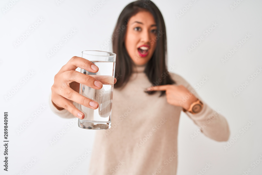 Young beautiful chinese woman holding glass of water standing over isolated white background with surprise face pointing finger to himself Young beautiful chinese woman holding glass of water standing over isolated white background with surprise face pointing finger to himself