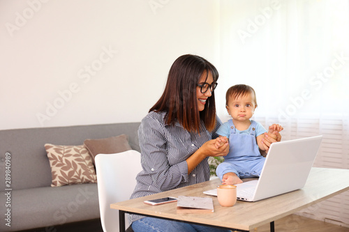 Stay at home mom working remotely on laptop while taking care of her baby. Young mother on maternity leave trying to freelance by the desk with toddler child. Close up, copy space, background. © Evrymmnt