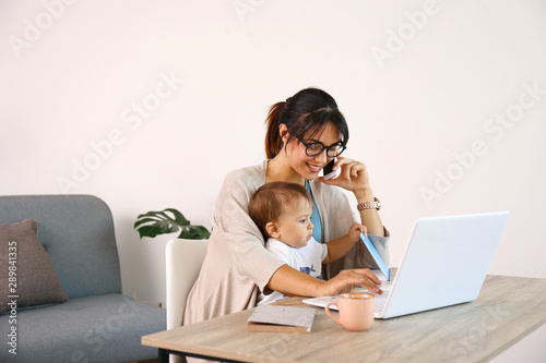 Stay at home mom working remotely on laptop while taking care of her baby. Young mother on maternity leave trying to freelance by the desk with toddler child. Close up, copy space, background.