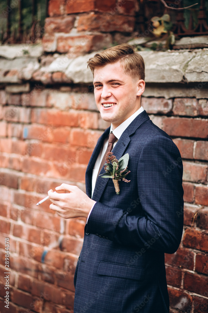 brutal groom in a suit and tie smokes a cigarette. The blond guy is walking around the old town. smoking, bad habits. Young male businessman on brick wall background. Portert of a smiling guy. brutal groom in a suit and tie smokes a cigarette. The blond guy is walking around the old town. smoking, bad habits. Young male businessman on brick wall background. Portert of a smiling guy.