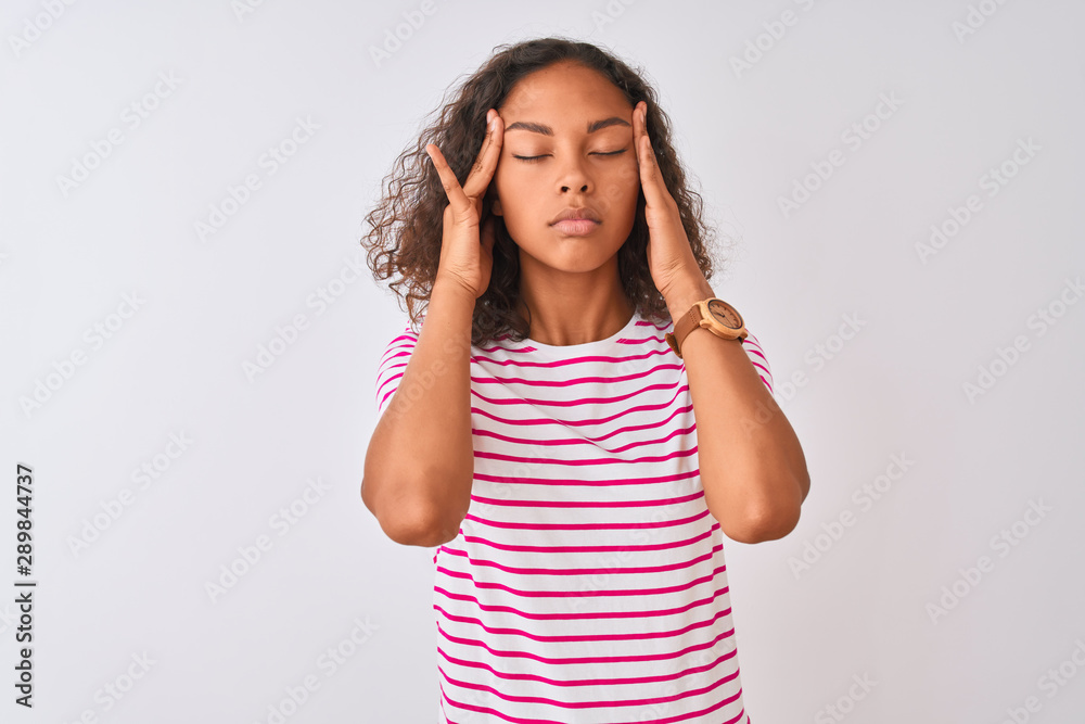 Young brazilian woman wearing pink striped t-shirt standing over isolated white background with hand on head for pain in head because stress. Suffering migraine. Young brazilian woman wearing pink striped t-shirt standing over isolated white background with hand on head for pain in head because stress. Suffering migraine.