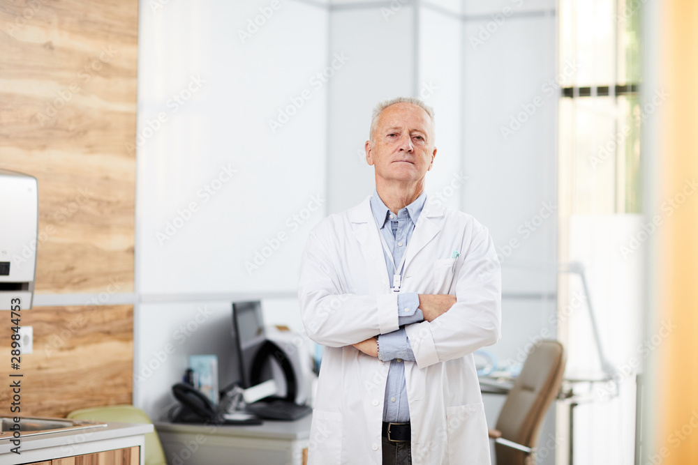 Portrait of experienced senior doctor wearing white coat posing in modern office standing with arms crossed and looking at camera, copy space