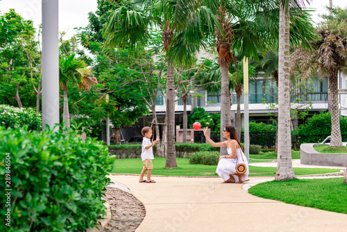 Full-length portrait of young woman and 4-year-old boy having a walk. Cute kid and his mother are playing ball. Tropical greenery in the background. Mom and son concept. Horizontal shot. Side view