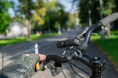 cigarette in the hand of a smoking cyclist who rides a bike on the background of the asphalted path of the city park