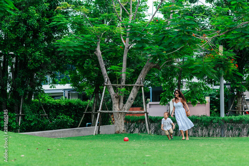 Full-length portrait of young woman and 4-year-old boy having a walk. Cute kid and his mother are playing ball. Tropical greenery in the background. Mom and son concept. Horizontal shot. Front view