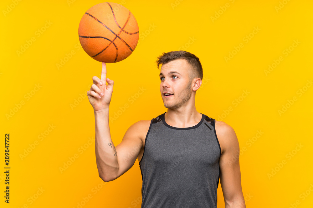 Young handsome blonde man holding a basket ball over isolated yellow background