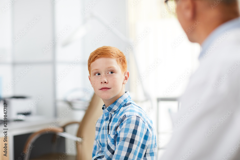Portrait of cute red haired kid looking at senior doctor during consultation in child healthcare clinic, copy space Portrait of cute red haired kid looking at senior doctor during consultation in child healthcare clinic, copy space