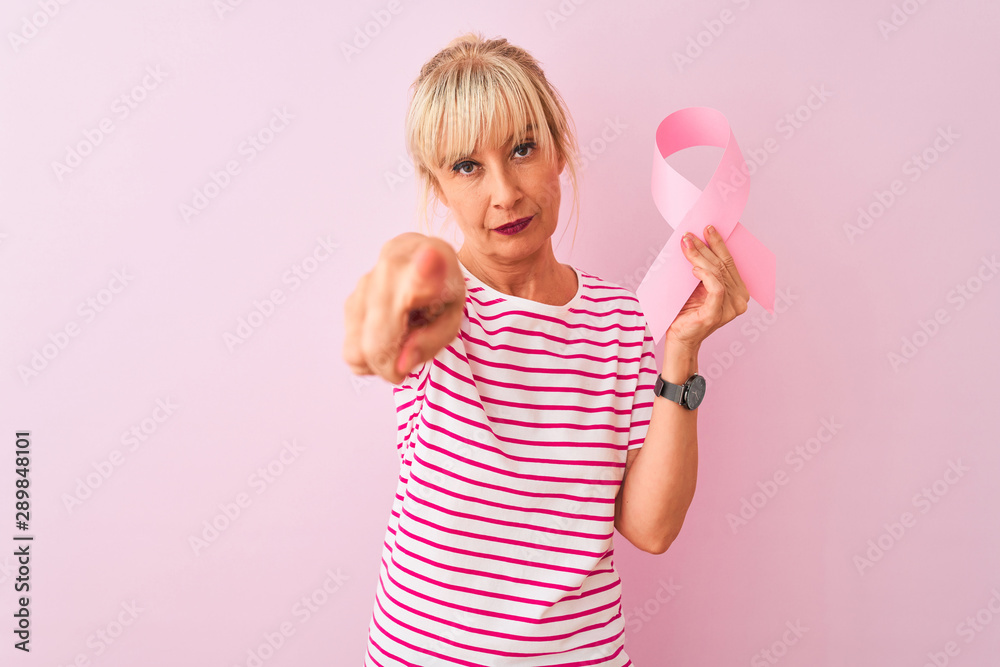 Middle age woman holding cancer ribbon standing over isolated pink background pointing with finger to the camera and to you, hand sign, positive and confident gesture from the front Middle age woman holding cancer ribbon standing over isolated pink background pointing with finger to the camera and to you, hand sign, positive and confident gesture from the front