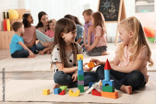 Cute girls playing with building blocks on floor while kindergarten teacher reading book to other children indoors photo