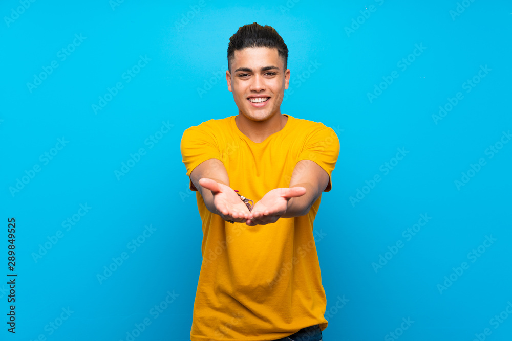 Young man with yellow shirt over isolated blue background holding copyspace imaginary on the palm to insert an ad Young man with yellow shirt over isolated blue background holding copyspace imaginary on the palm to insert an ad