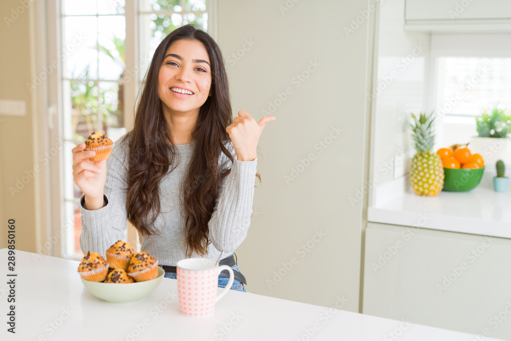 Young woman eating chocolate chips muffins and drinking coffee pointing and showing with thumb up to the side with happy face smiling Young woman eating chocolate chips muffins and drinking coffee pointing and showing with thumb up to the side with happy face smiling