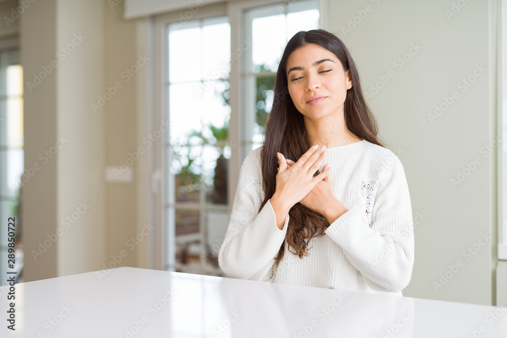 Young beautiful woman at home on white table smiling with hands on chest with closed eyes and grateful gesture on face. Health concept. Young beautiful woman at home on white table smiling with hands on chest with closed eyes and grateful gesture on face. Health concept.