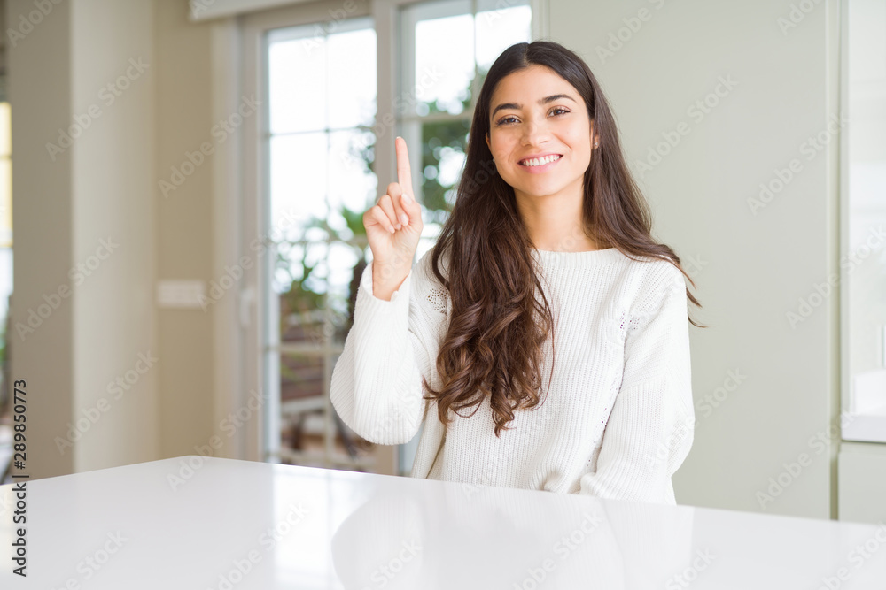 Young beautiful woman at home on white table showing and pointing up with finger number one while smiling confident and happy. Young beautiful woman at home on white table showing and pointing up with finger number one while smiling confident and happy.