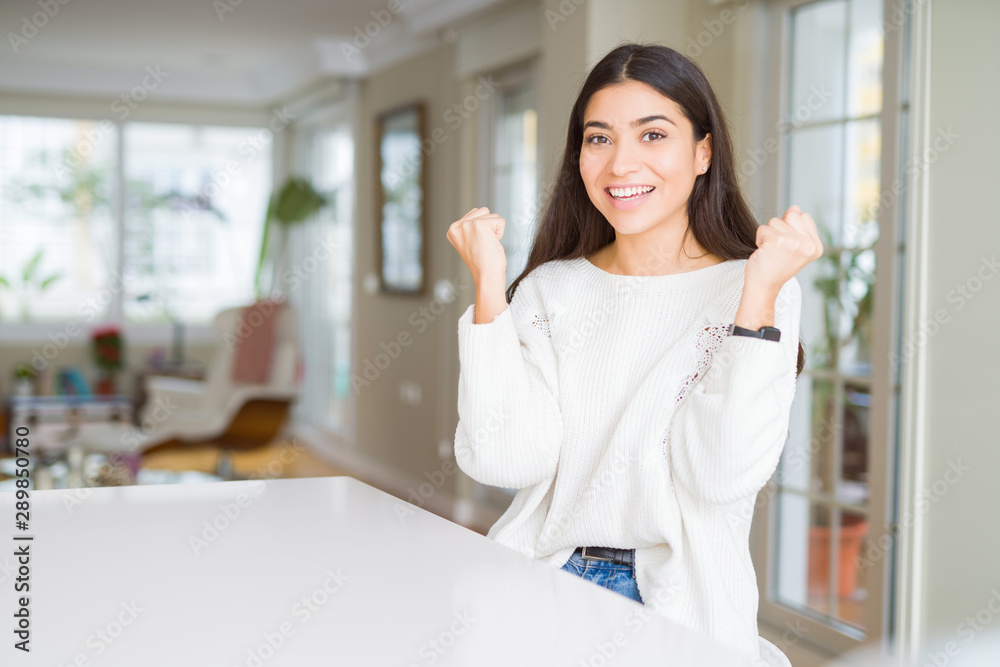 Young beautiful woman at home on white table celebrating surprised and amazed for success with arms raised and open eyes. Winner concept. Young beautiful woman at home on white table celebrating surprised and amazed for success with arms raised and open eyes. Winner concept.