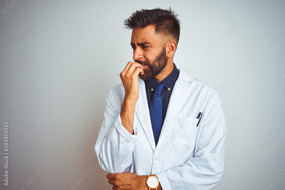 Young indian doctor man standing over isolated white background looking stressed and nervous with hands on mouth biting nails. Anxiety problem. Young indian doctor man standing over isolated white background looking stressed and nervous with hands on mouth biting nails. Anxiety problem.