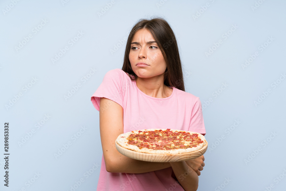 Pretty young girl holding a pizza over isolated blue wall making doubts gesture while lifting the shoulders Pretty young girl holding a pizza over isolated blue wall making doubts gesture while lifting the shoulders