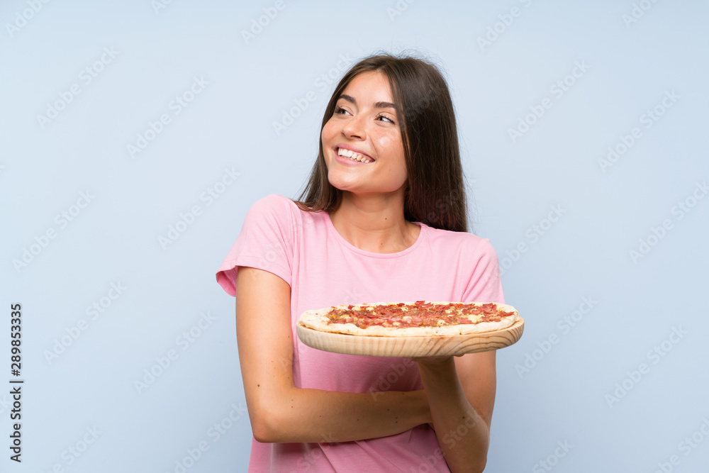 Pretty young girl holding a pizza over isolated blue wall looking up while smiling Pretty young girl holding a pizza over isolated blue wall looking up while smiling