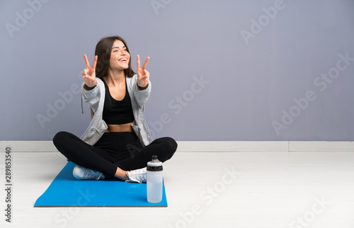 Young sport woman sitting on the floor with mat smiling and showing victory sign